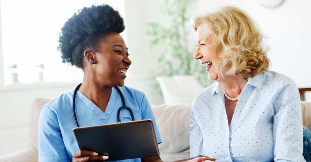 BIPOC healthcare professional holding a mobile tablet talks with elderly blond patient