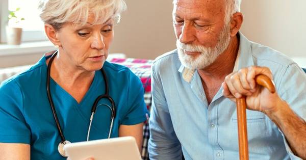 Healthcare professional in scrubs shows information on a tablet to an elderly patient with a cane