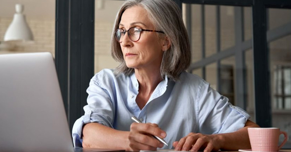 woman with gray hair writes while looking at laptop