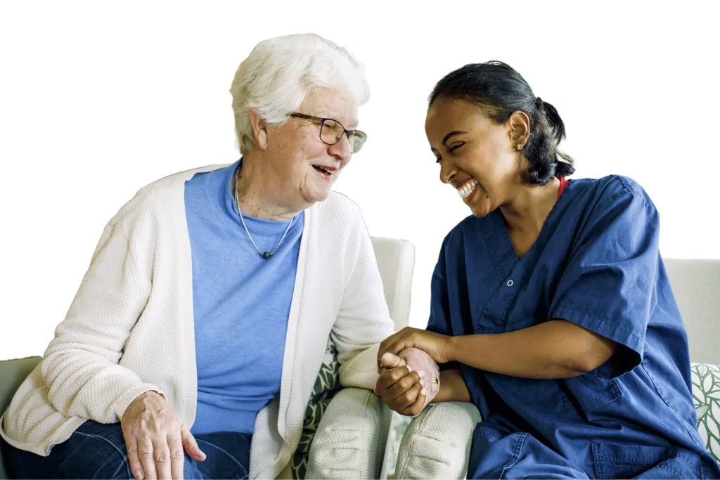 Smiling nurse in navy scrubs holds hands with an older woman as they talk on a sofa