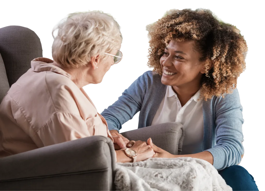 Caregiver and an older woman smile and hold hands while talking in a living room